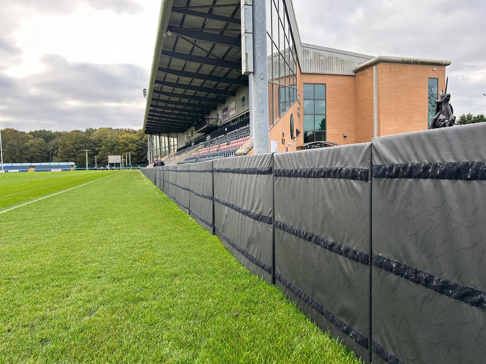 Grey padded barrier runs along the green pitch toward a covered spectator stand and brick building with glass windows and an equestrian statue.